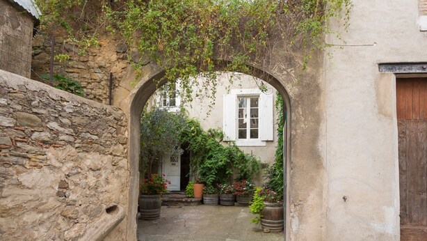 Un portail en arc donne sur une cour bordée de murs en pierre, agrémentée de plantes en pot et surmontée d’un bâtiment blanc doté d’une porte en bois.
