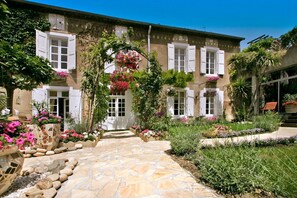 A stone pathway leading to a building with white shutters and a garden with flowers and plants.