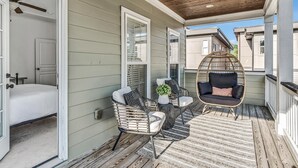 A covered porch with a bed, two wicker chairs, a round wicker chair, and a small table with a plant.