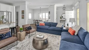 A modern living room with a blue sofa, a brown leather chair, and a round coffee table. There is a kitchen area with white cabinets and a stainless steel refrigerator.