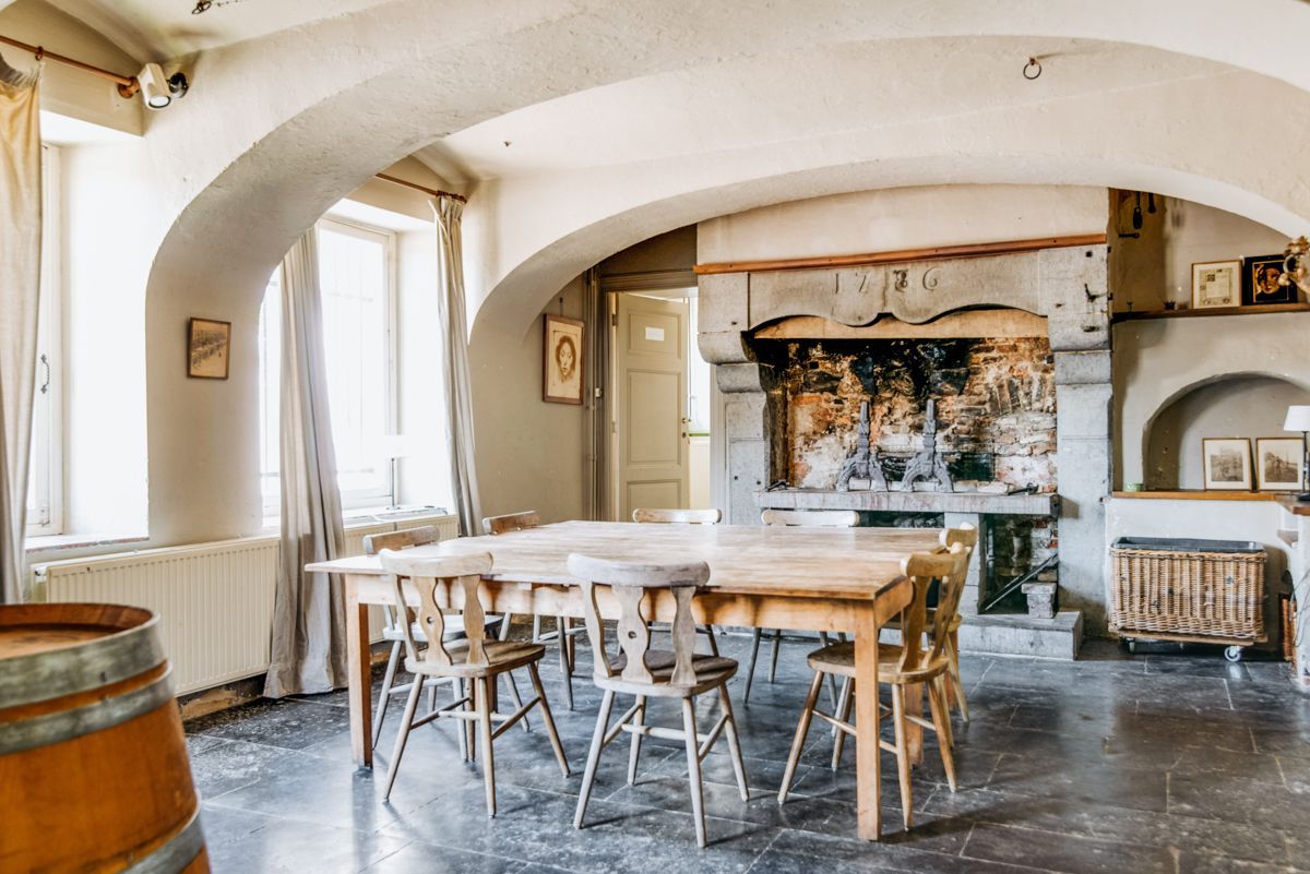 Un espace repas comprenant une longue table et des chaises en bois, une cheminée en pierre et une vue sur l’extérieur grâce à une fenêtre en arc de cercle.