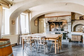 A dining area with a long wooden table and chairs, a stone fireplace, and a view of the outdoors through an arched window. - Chateau Alois - Fourteen Bedroom Castle, Sleeps 35 (Ciney)