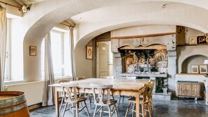 A dining area with a long wooden table and chairs, a stone fireplace, and a view of the outdoors through an arched window.