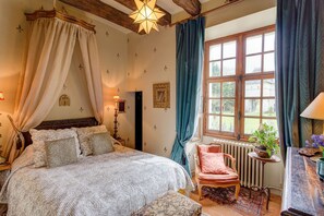 A bedroom with a four-poster bed, a chair, a table with a plant, and a window with curtains.