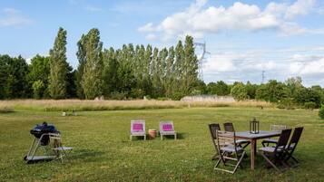 Een terras buiten met een tafel, stoelen, een barbecue en een picknicktafel met kussens.