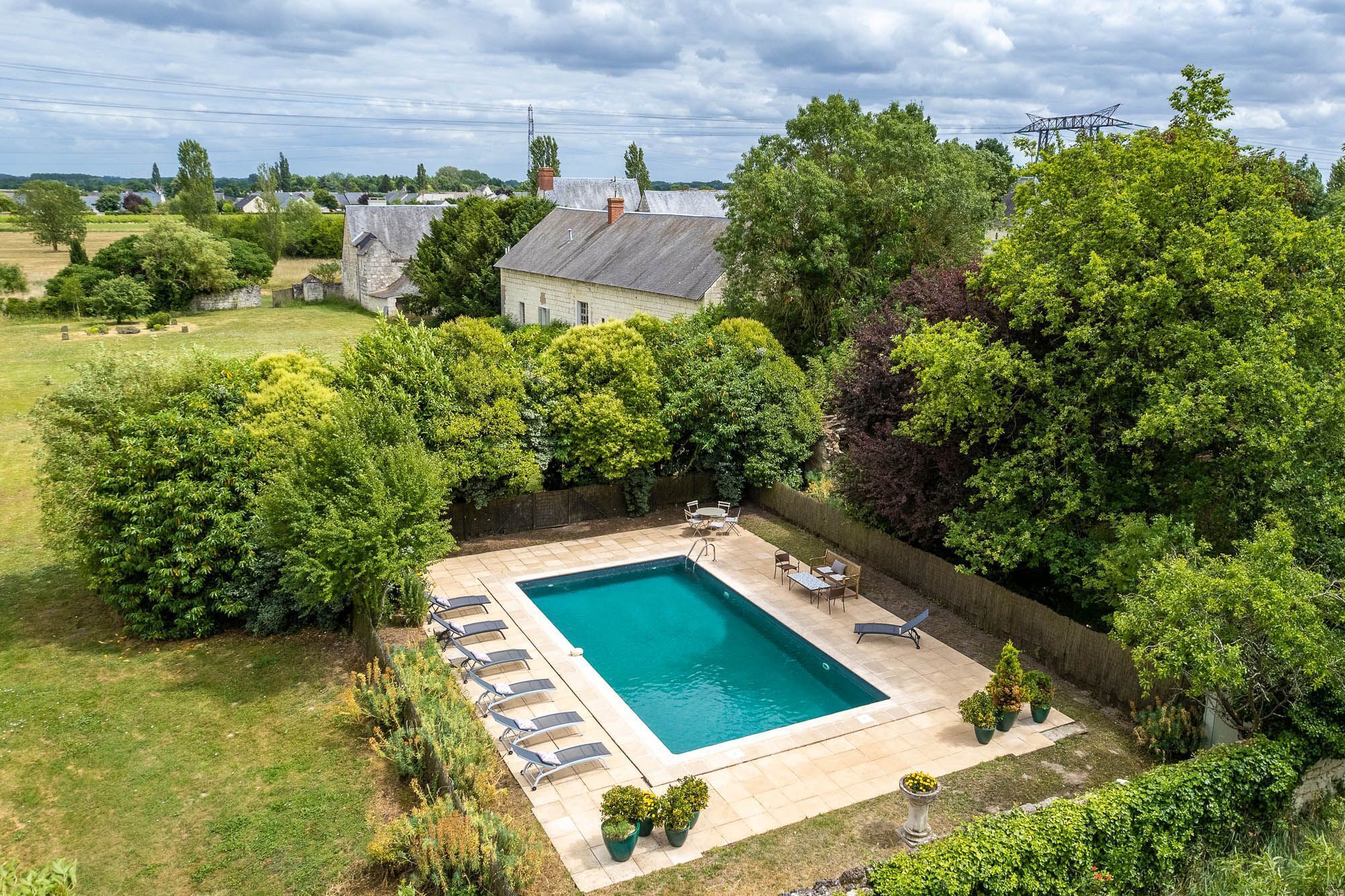 A rectangular swimming pool with a tiled deck, surrounded by lush greenery and trees.