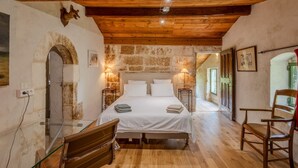 A rustic bedroom with a stone wall, wooden ceiling beams, a bed with white linens, a glass table, and a wooden chair.