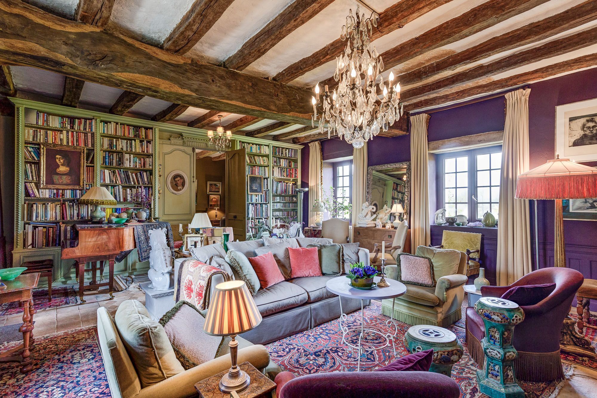 Ein Zimmer mit freiliegenden Holzbalken, einer Kristallleuchter, einem Kamin und einer großen Anzahl an Bücherregalen.