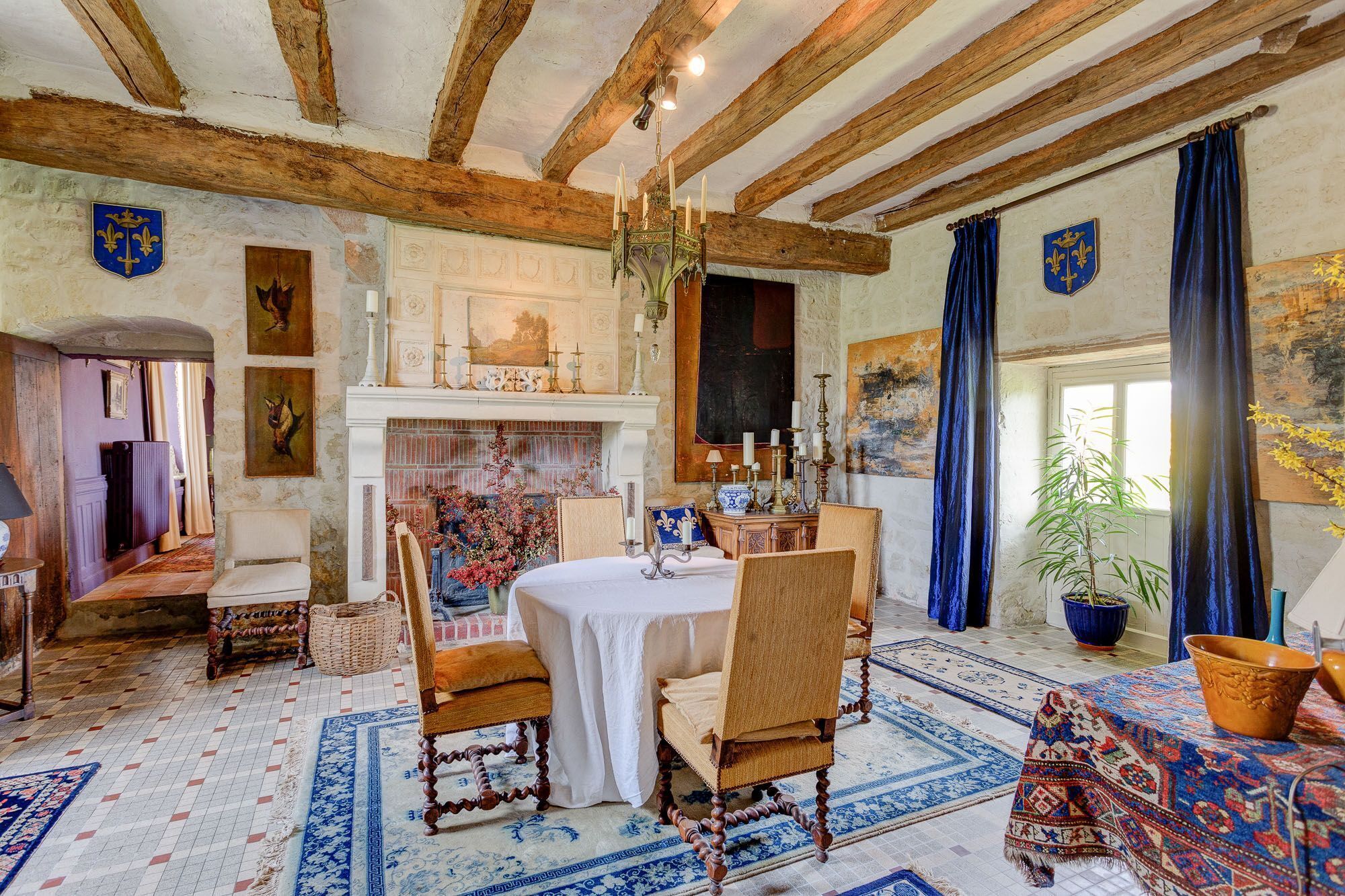 Een klassieke kamer met zichtbare houten balken, een open haard, een eettafel met stoelen en een groot raam met gordijnen.