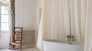 Une salle de bain avec une baignoire, une chaise en bois, un radiateur et une fenêtre avec des rideaux.