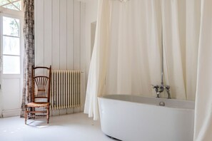 A bathroom with a bathtub, a wooden chair, a radiator, and a window with curtains.