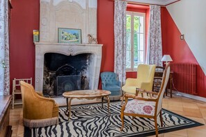 Een kamer met een open haard, twee fauteuils, een tafel en een raam met gordijnen.