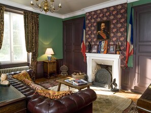 Une pièce à l’ambiance traditionnelle, avec une cheminée, un canapé, une table avec des livres et un portrait au-dessus du manteau de la cheminée.