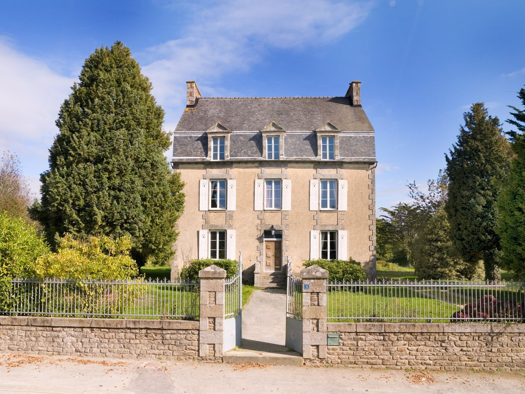 A two-story stone building with a symmetrical facade, a central entrance with steps, and a gated driveway.