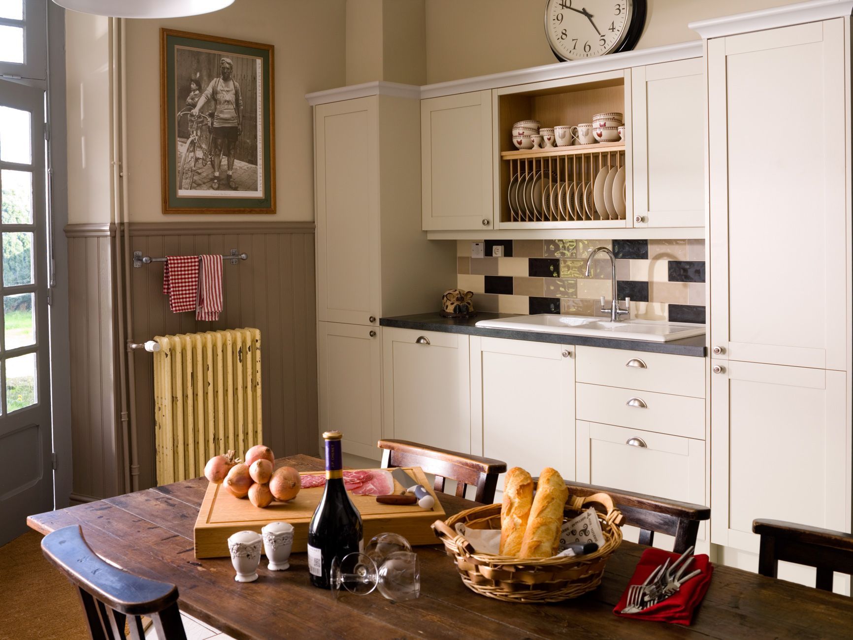 A kitchen with white cabinets, a clock, a framed picture, and a dining table with bread, wine, and utensils.