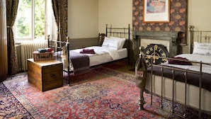 A traditional bedroom with two beds, a fireplace, a patterned carpet, and a window with curtains.