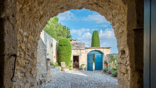 Un patio empedrado con un arco de piedra, una puerta azul y un alto árbol con forma cónica.