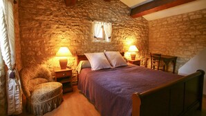 Ein Schlafzimmer mit einer Steinwand, einem hölzernen Bettgestell, zwei Nachttischlampen, einem kleinen runden Hocker und einem Fenster mit Vorhängen.
