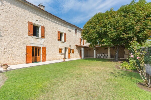 A stone building with wooden shutters, a grassy courtyard, and a tree.