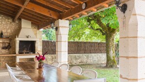 Un patio al aire libre con una mesa de madera y sillas blancas, una chimenea de piedra y vistas a un árbol y jardín.