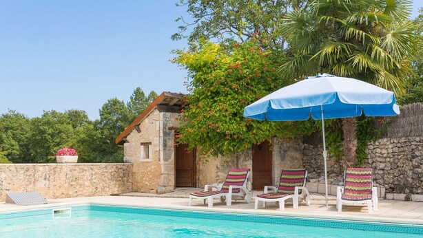 Zona de piscina con un edificio de piedra, una sombrilla azul y sillones de relax coloridos.