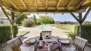 Una pérgola de madera con una mesa lista para comer, con vista a una piscina y un jardín ajardinado.