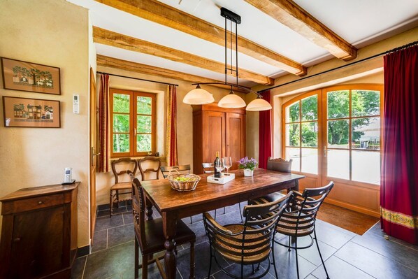 Un comedor con una mesa de madera, cuatro sillas, una araña y ventanas grandes con cortinas.