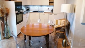 Een moderne keuken met witte kasten, een houten eettafel met transparante stoelen, en een rieten stoel.