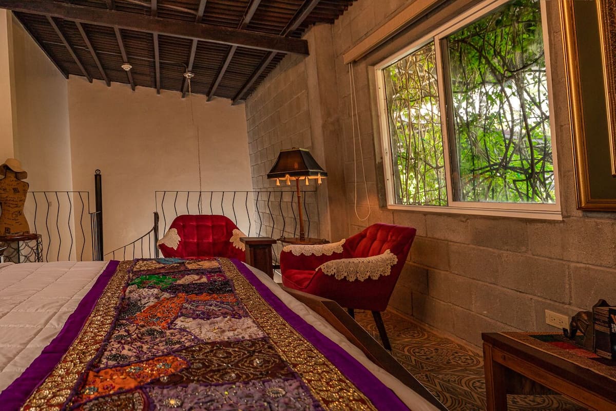 A bedroom with a large bed, a red chair, a window with a view of trees, and a wall with a decorative piece.