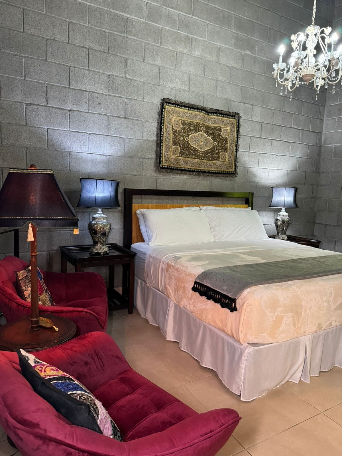 Un dormitorio con pared de ladrillo, una cama con ropa de cama blanca, una silla roja, dos mesitas de noche con lámparas y una araña.