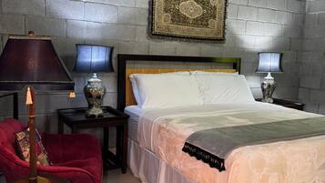 Un dormitorio con pared de ladrillo, una cama con ropa de cama blanca, una silla roja, dos mesitas de noche con lámparas y una araña.