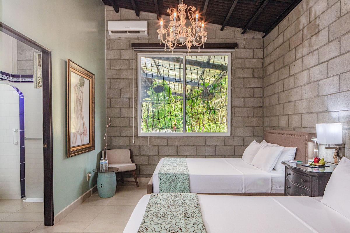 A hotel room with two beds, a chandelier, a window with a view of greenery, a framed picture on the wall, and a small table with a bottle.