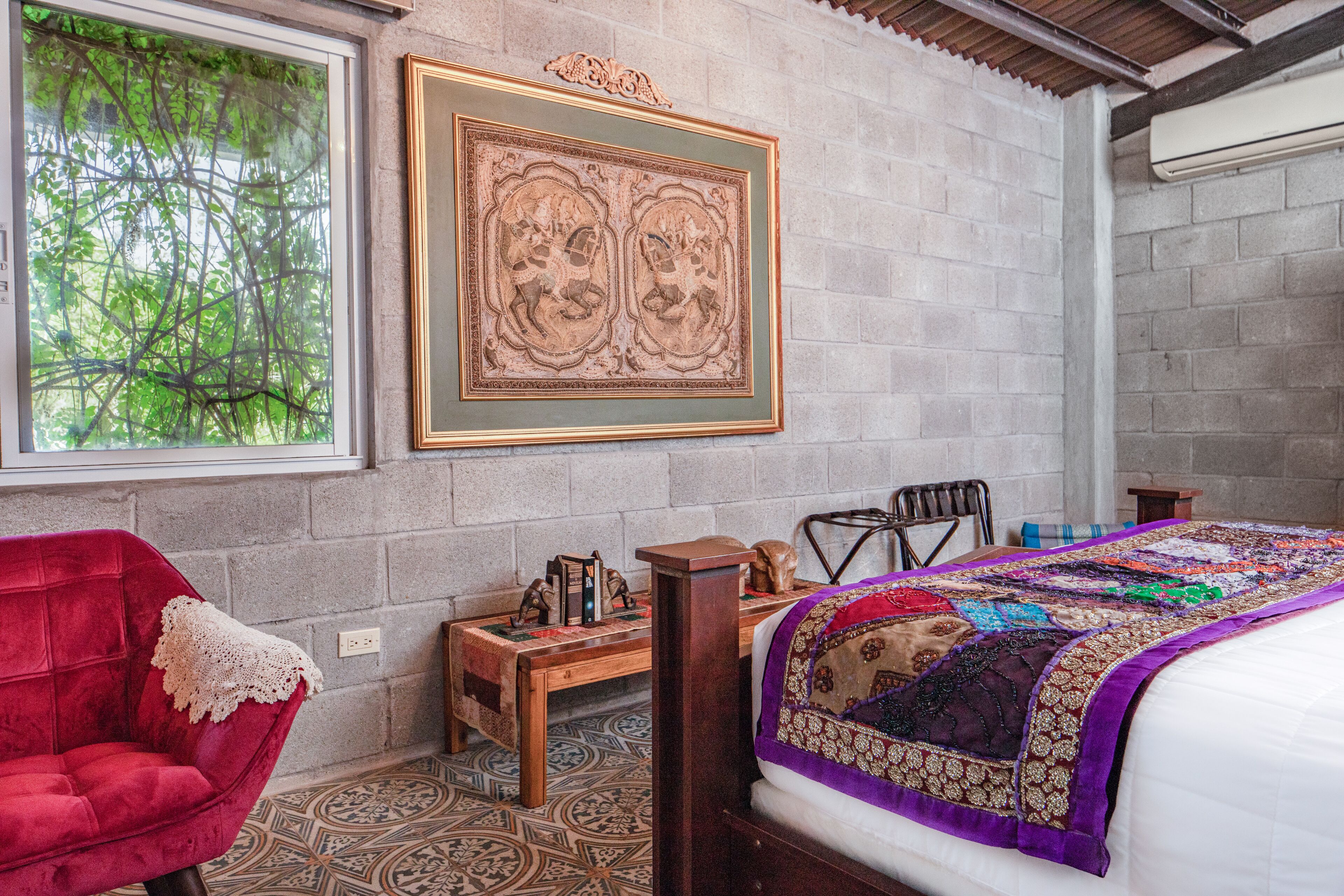 Une chambre avec une grande fenêtre, un fauteuil rouge, une table de chevet en bois, un lit recouvert d’une courtepointe colorée et une œuvre d’art encadrée accrochée au mur.