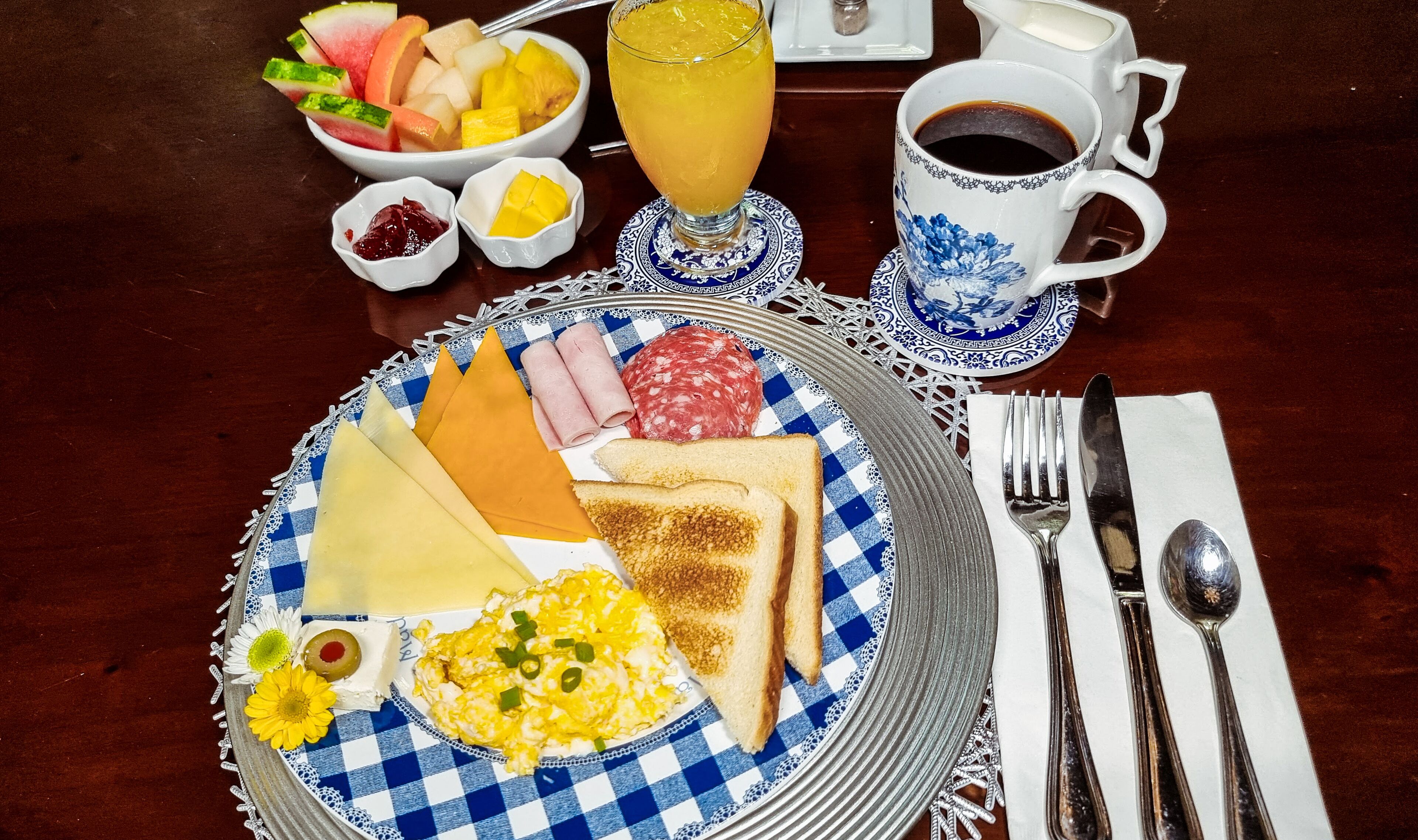 Ein Frühstückstisch mit Toast, Eiern, Käse, Schinken, Salami und einer Obstschale. Dazu eine Tasse Kaffee und ein Glas Orangensaft.