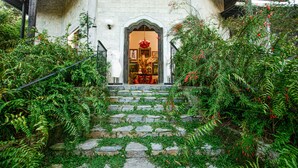 Una escalera de piedra conduce a la entrada de un edificio con un arco tradicional, rodeada de exuberante follaje y flores rojas.