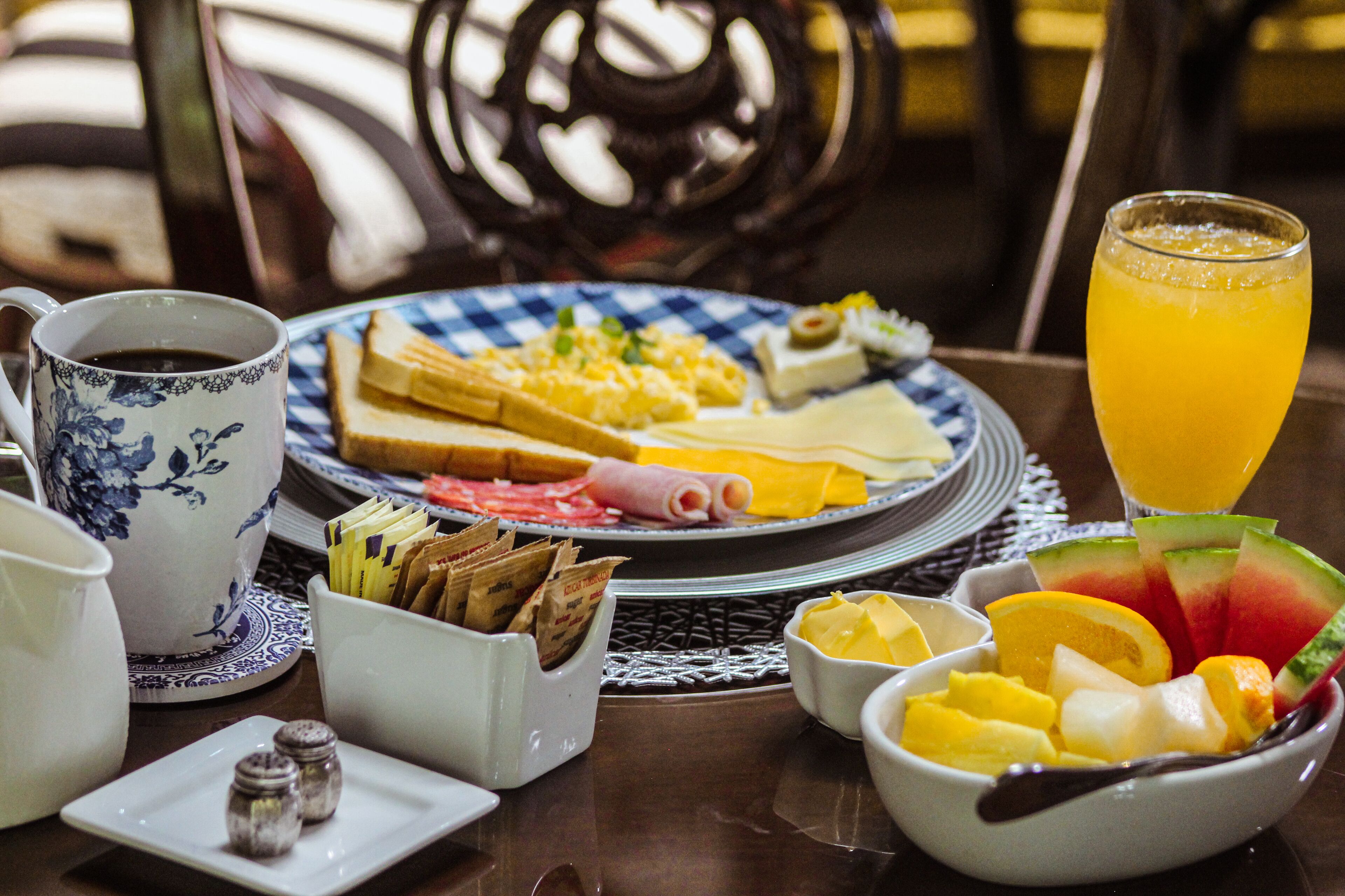 A breakfast table with a plate of eggs, ham, cheese, and toast, a bowl of fruit, a glass of orange juice, and a coffee cup.