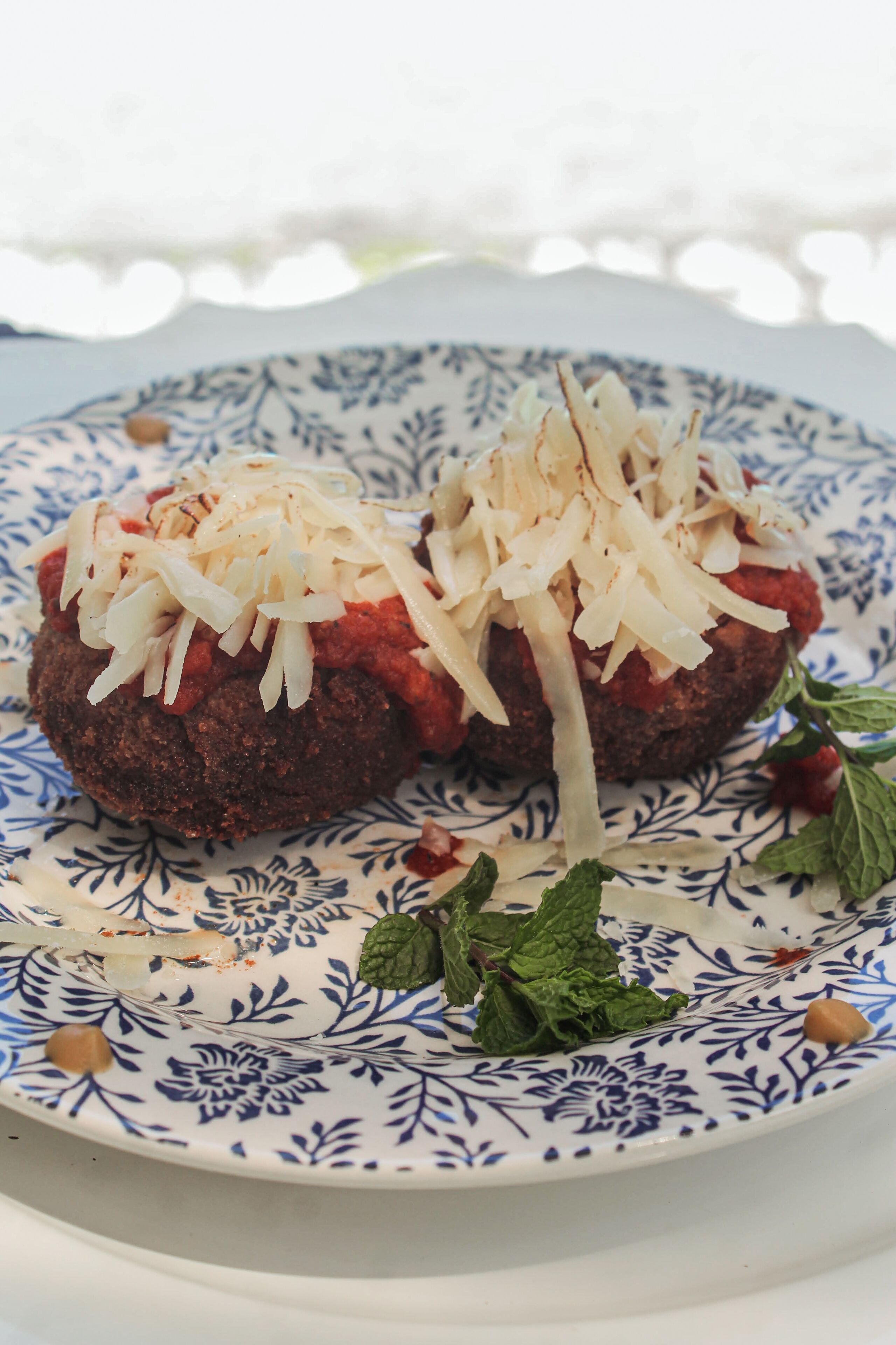 A plate with two meatballs topped with shredded cheese and tomato sauce, garnished with mint leaves.