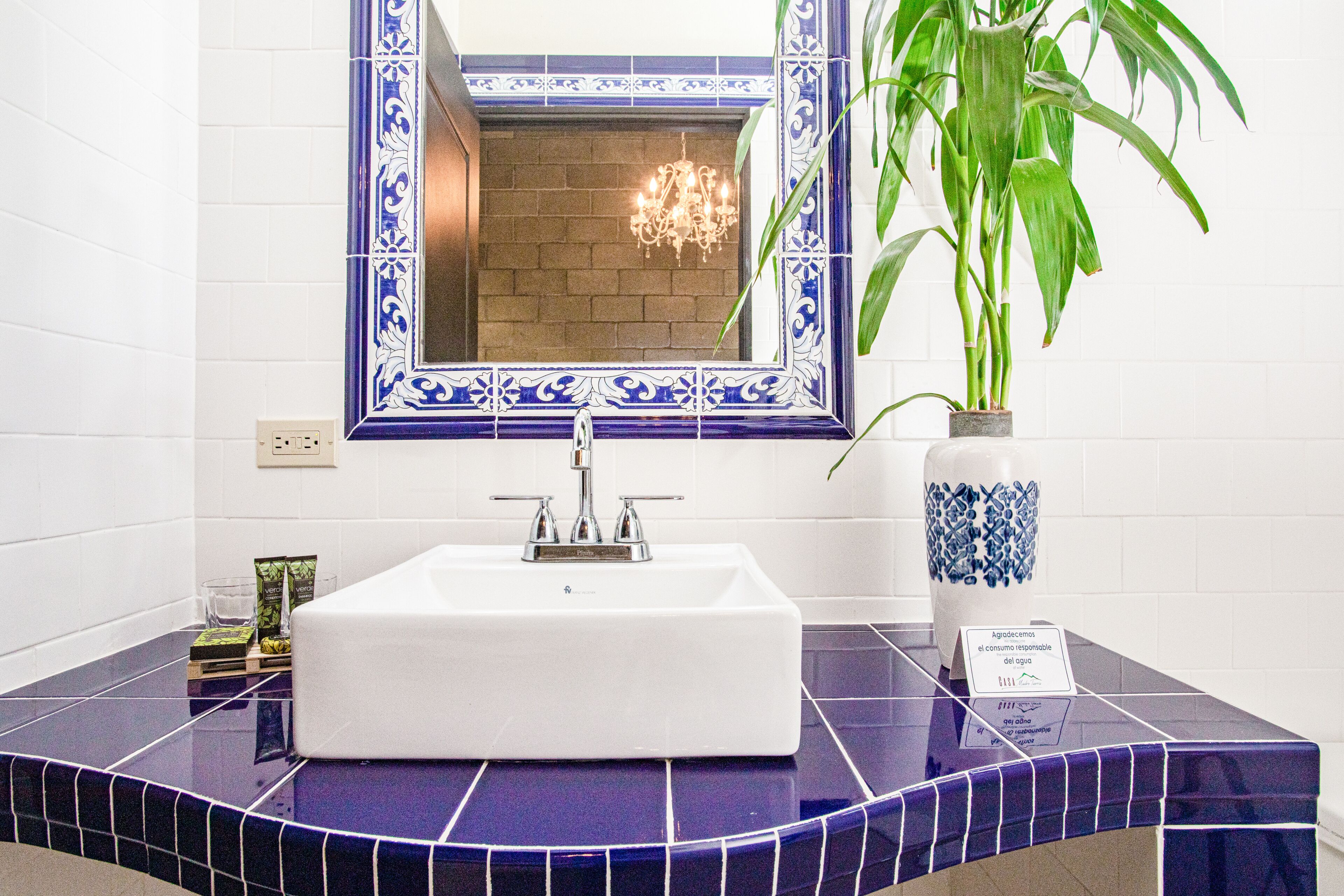 A bathroom with a white sink, blue and white tiles, a potted plant, and a mirror with a blue frame.