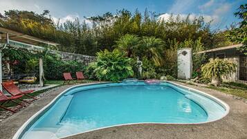 A swimming pool with red lounge chairs, surrounded by lush greenery and a stone wall.