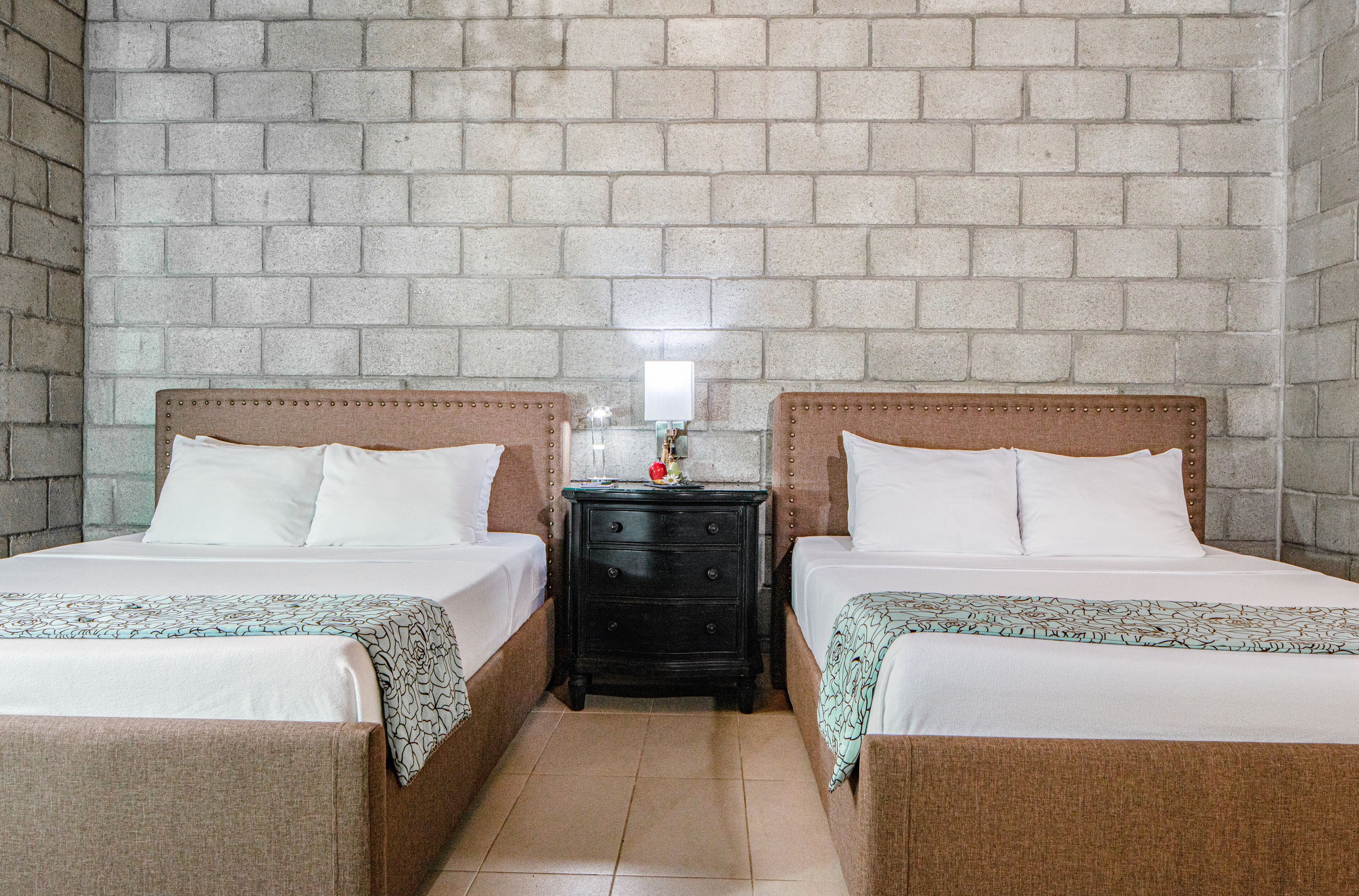 A room with two beds, a nightstand with a lamp, and a brick wall.
