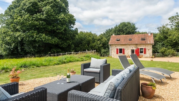 A patio with wicker furniture, a stone house with red shutters, and a large tree in the background.