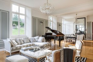 A spacious living room with a grand piano, a white sofa set, and a chandelier. - Chateau Quatre Saisons (Domjean)