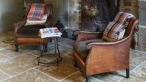 A rustic room with a stone fireplace, two leather armchairs, a small table with a magazine, and a plaid throw blanket on one chair. - Chateau Quatre Saisons (Domjean)