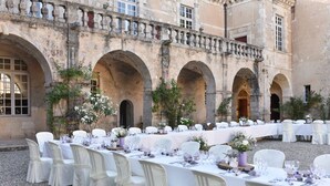 Une salle de banquet en plein air, avec des tables et des chaises recouvertes de nappes blanches, située devant un bâtiment historique orné d’arcades.