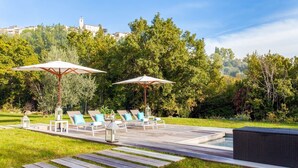A poolside area with lounge chairs and umbrellas, surrounded by greenery and a distant building.