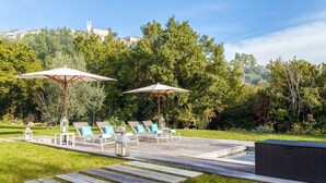 Een terras met loungezetels en parasols, een zwembad en uitzicht op een heuvel met bomen en gebouwen.