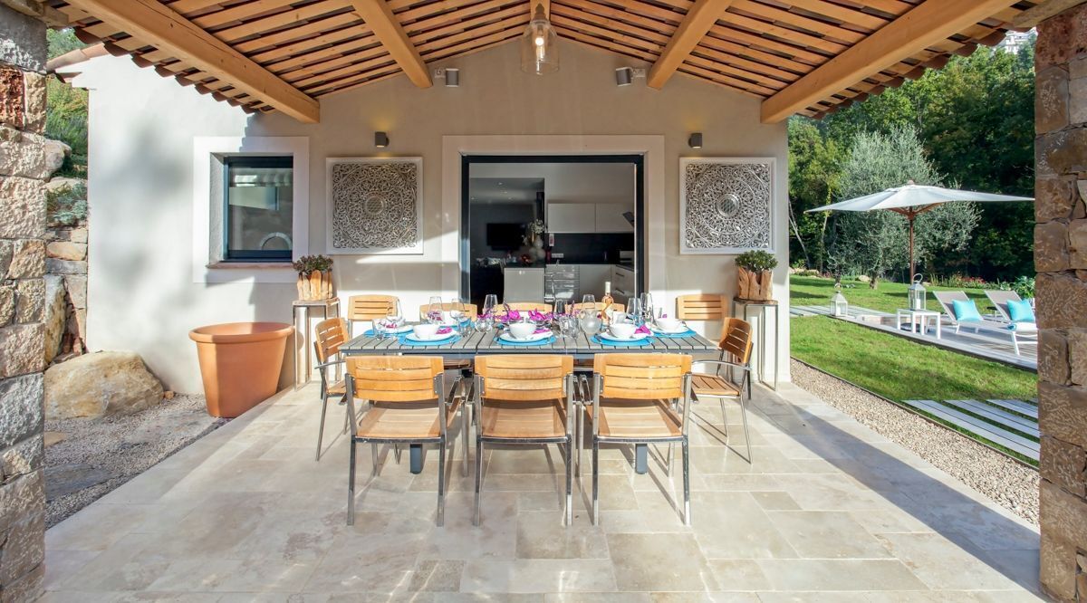 An outdoor patio with a table set for dining, surrounded by stone walls and a wooden pergola.