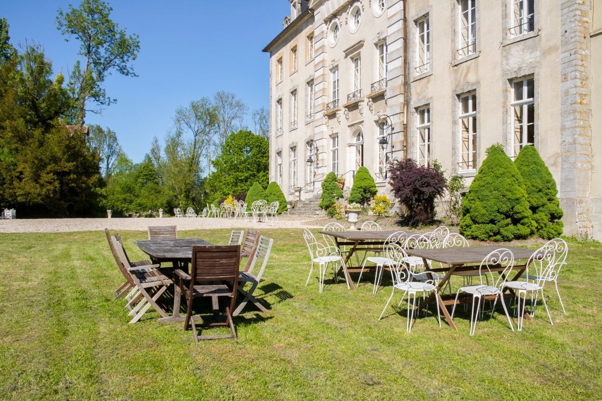 An outdoor dining area with tables and chairs, a building with classical architecture in the background, and a well-maintained lawn.