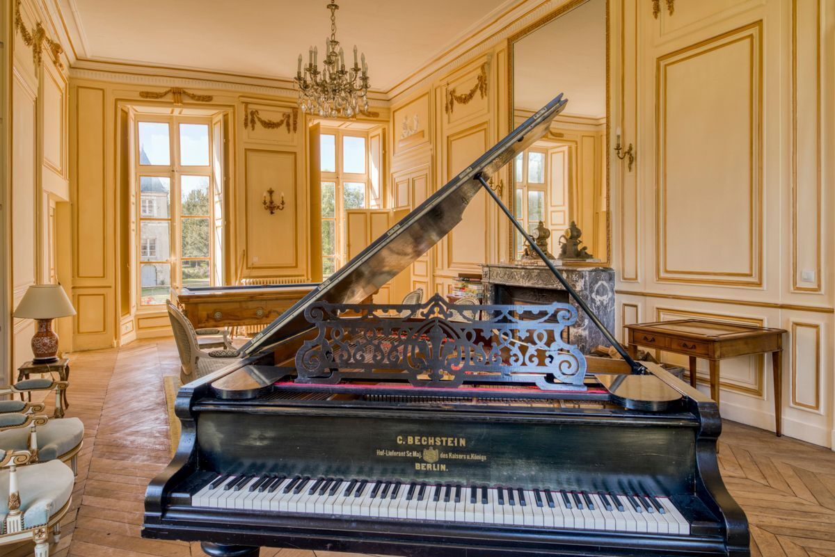 A grand piano in a spacious room with ornate wall paneling and a chandelier.