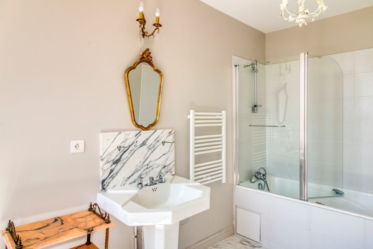 Une salle de bain avec un lavabo blanc, un pare-chocs en marbre, un miroir avec un cadre doré, une douche en verre et un meuble-lavabo en bois.
